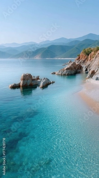 Fototapeta Vibrant Turquoise Ocean Water Shimmers Under Bright Daylight Sunlight on a Rocky Beach Shoreline with Lush Green Distant Hills and Majestic Mountains Under a Clear Blue Sky
