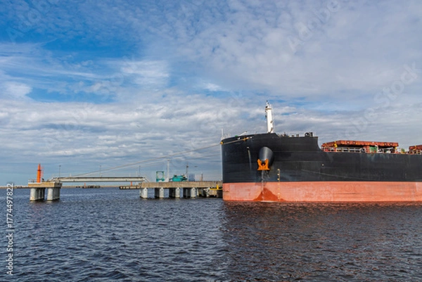 Fototapeta Large cargo ship docked at an industrial port terminal. Mooring cables are attached to the ship. Industrial port equipment and pipelines are visible in the background