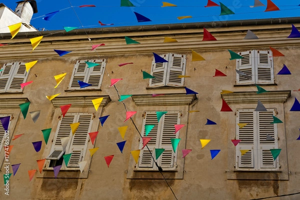 Fototapeta Multicolored pennant flags strung across a beige building with white window shutters under a deep blue sky, creating a festive street atmosphere.