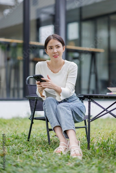 Fototapeta Asian woman looking at smartphone in hand writing in notebook and sitting at wooden table. Study or Working Outdoor.

