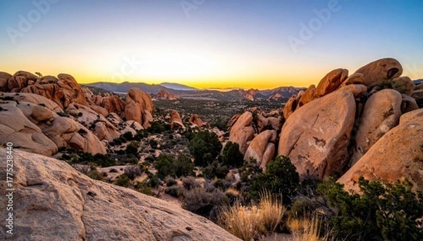 Fototapeta Rugged Desert Landscape at Dusk Featuring Large Rock Formations and Distant Town Lights Under a Warm Hued Sky