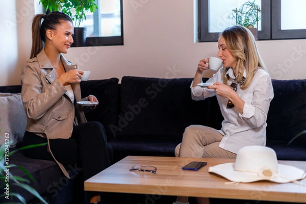 Fototapeta Two women enjoy coffee together in a cozy seating area with plants in the background