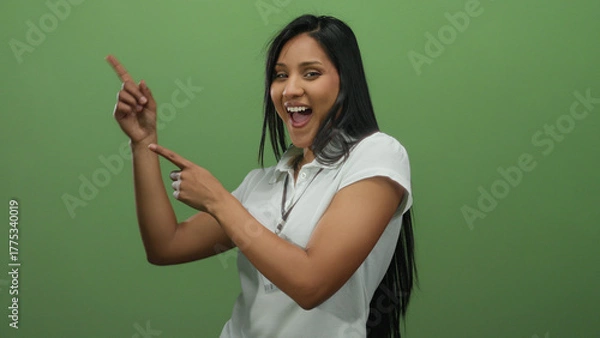 Fototapeta Woman smiling and pointing sideways wearing a white shirt with a badge against a green wall, exuding a friendly and inviting atmosphere.