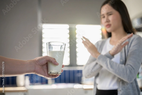 Fototapeta Allergy dairy food lactose intolerance allergen health care illness concept. Selective focus on a glass of milk with blurred woman gesturing refusing stop or reject say no to cow milk.