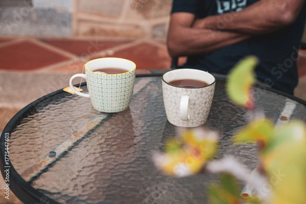 Fototapeta Close-up of two ceramic cups with morning tea on glass table with cropped image of a South Asian man sitting hand-crossed. Selective focus on the cup.