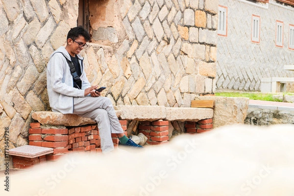 Fototapeta Tourist sitting on a bench smiling and browsing phone in front of a Fujian-style house in Kinmen. Exploring travelling Island of Taiwan, selective focus.