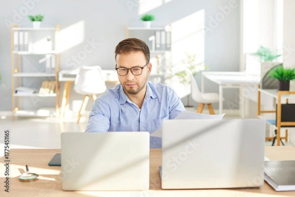 Fototapeta Confident young attractive business man in formal shirt and glasses looking seriously at computer monitor screen working at the desk on his workplace and typing on laptop in office.