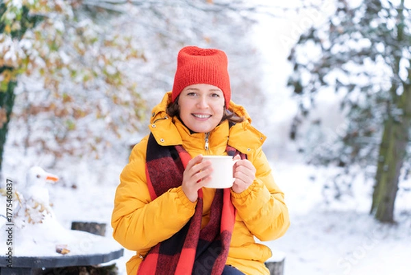 Fototapeta Smiling woman in red hat and yellow jacket enjoys hot drink from white mug in snowy park, radiating warmth and joy during cold winter. Self-care and happiness in outdoor leisure. Holiday warmth