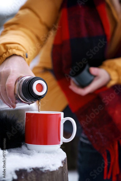 Fototapeta Close-up of senior woman hands pouring hot tea from vacuum flask into red mug outdoors in snowy winter setting. Outdoor comfort and warmth in winter. Simple pleasures as warming drink in cold weather