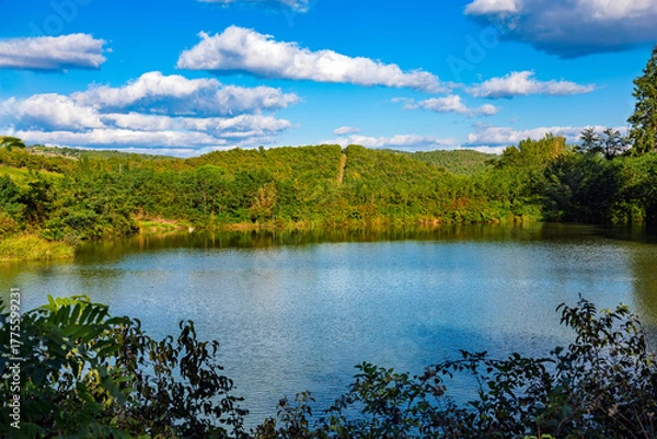 Obraz 2025-10-02 CALM LAKE FRAMED AND SURROUNDED BY GREEN FOLIAGE AND A ROLLING HILL IN THE BACKGROUND AND A NICE CLOUDY SKY NEAR SIENA IN THE TUSCUNY REGION OF ITALY