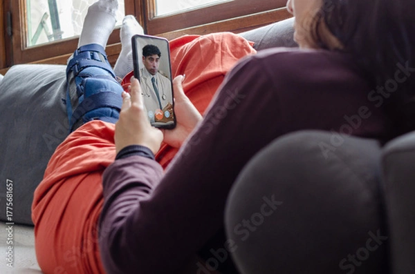 Fototapeta Young woman with broken leg laying on a couch having a video call with a doctor.