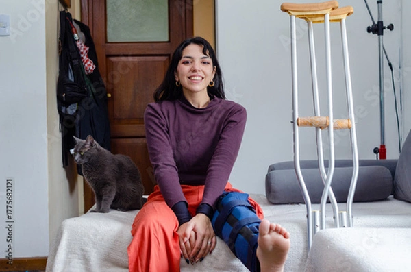 Fototapeta Portrait of a smiling young woman with a broken leg sitting on a couch.