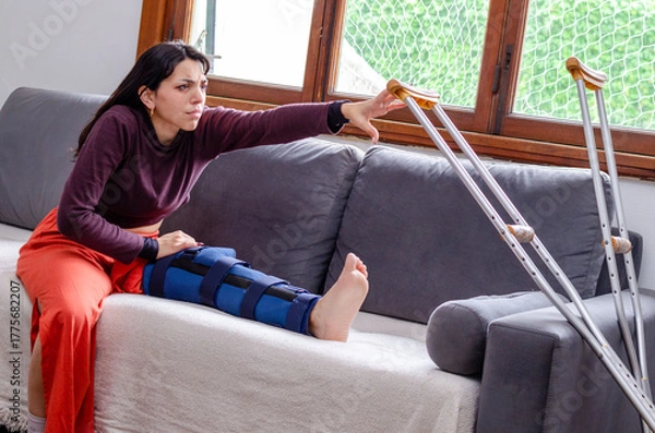 Fototapeta Young woman with a broken leg trying to reach for a crutch.