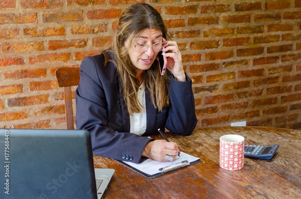Fototapeta Mature woman working with a laptop and having a conversation on phone.Woman taking notes while talking on mobile phone.