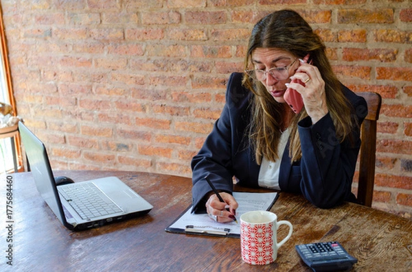 Fototapeta Mature woman working with a laptop and having a conversation on phone.Woman taking notes while talking on mobile phone.