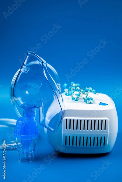 Fototapeta Close-up side view of a blue mask and a device on a blue background and flowers nearby. The concept of treating respiratory diseases, lung diseases, prevention and self-care. High quality photo