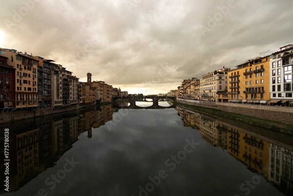 Fototapeta View of the Arno River in Florence, Tuscany, Italy, with a stone bridge and colorful historic buildings reflected in the calm water under a dramatic cloudy sky. Classic Italian cityscape.