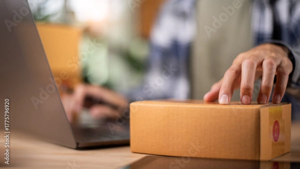 Fototapeta A small business owner prepares a cardboard box for delivery while managing online orders on a laptop, symbolizing e-commerce, logistics, and digital retail efficiency.