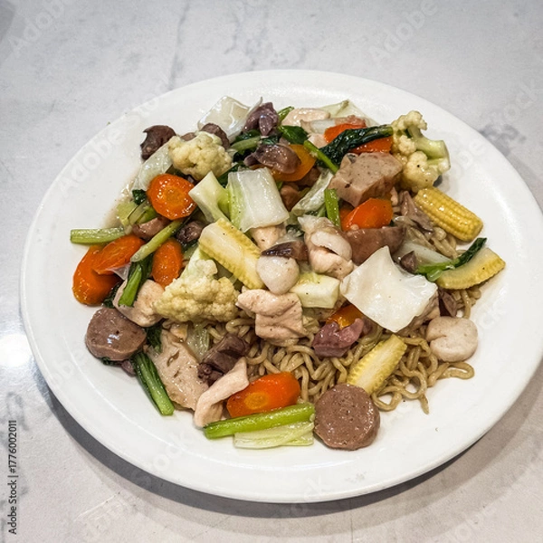 Fototapeta Stir-fried noodles topped with mixed vegetables, chicken, and meatballs, served on a white plate. Colorful and appetizing Asian cuisine.