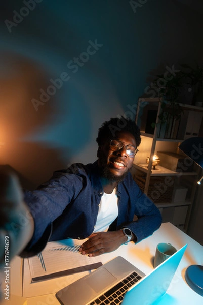 Obraz Vertical selfie of happy young African American man looking at camera while studying late at night at home office.