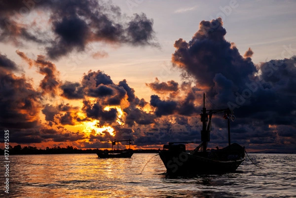 Fototapeta silhouette of two fishing boats is set against the shimmering water as the sun dips behind massive, dark, and intense orange clouds. The atmosphere is powerful and evocative of twilight.
