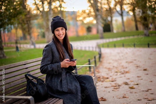 Obraz Confident smiling woman sitting at the city park and using a smartphone