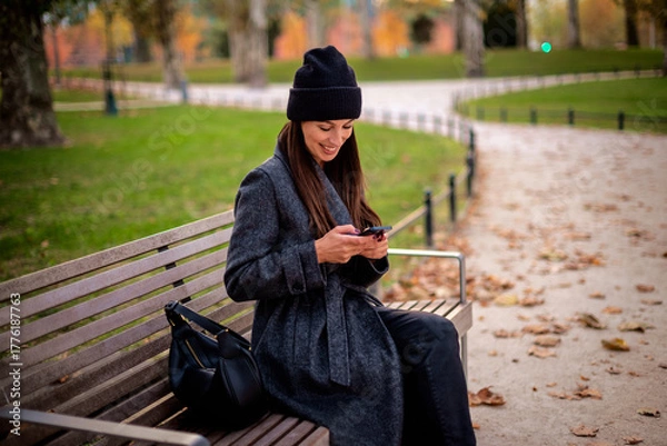 Obraz Confident smiling woman sitting at the city park and using a smartphone