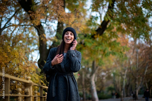 Obraz Stylish middle-aged woman wearing long coat and knitted beanie and using smartphone outdoors