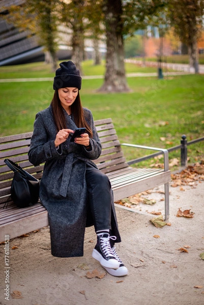 Obraz Confident smiling woman sitting at the city park and using a smartphone