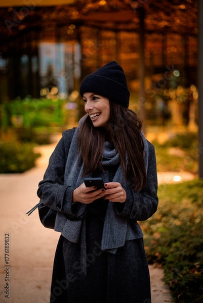 Obraz Happy woman wearing beanie and knitted scarf and using her smartphone on the street