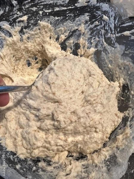 Fototapeta Thick, sticky bread dough with visible grains being mixed in a glass bowl using a metal spoon