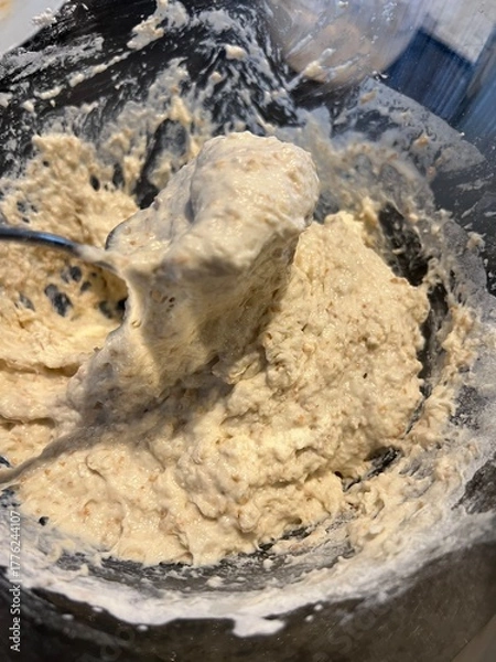 Fototapeta Thick, sticky bread dough with visible grains being mixed in a glass bowl using a metal spoon