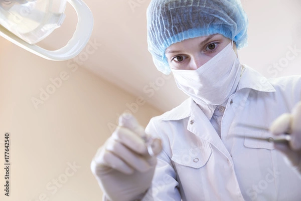 Fototapeta female dentist in medical mask, cap and gloves with a concentrated expression on his face holds the dental tools. Portrait with the lower angle