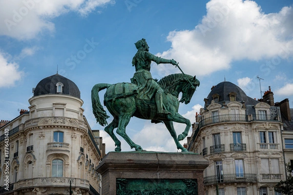 Fototapeta Bronze statue of Joan of Arc prominently framed between two historic buildings in Orléans, capturing the heroic figure against the urban backdrop under clear daylight.