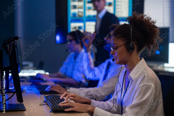 Fototapeta Diverse team of professionals customer service agent wearing headsets and monitoring computer working in dark control room. Female support and assistance call center at night in modern office.