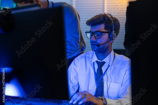 Fototapeta Man support and assistance call center at night in modern office. Diverse team of professionals customer service agent wearing headsets and monitoring computer working in dark control room.