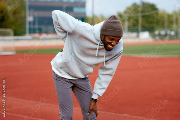 Fototapeta Male athlete cries out in pain, holding his lower back with his hand due to a serious injury or muscle cramp during a break in his outdoor workout