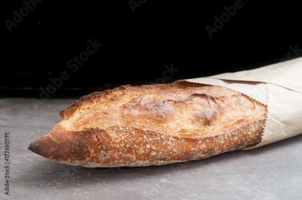Obraz Rustic sourdough baguette served wrapped in paper on a grey slate counter top.