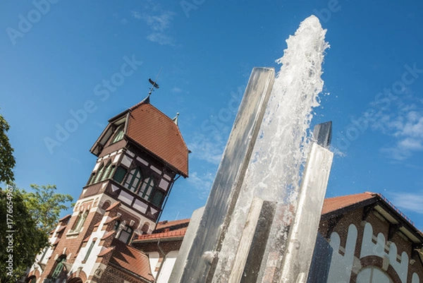 Fototapeta Moderner Springbrunnen aus poliertem Metall mit Wasserfontäne vor denkmalgeschützter alter Turnhalle mit Fachwerk und Walmdach in Metzingen. 