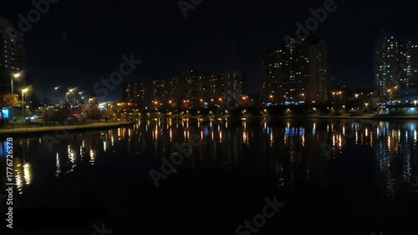 Fototapeta Night cityscape with lights reflecting on lake. Tall residential buildings illuminated at night with colorful streetlights reflecting on a calm urban lake in a peaceful setting