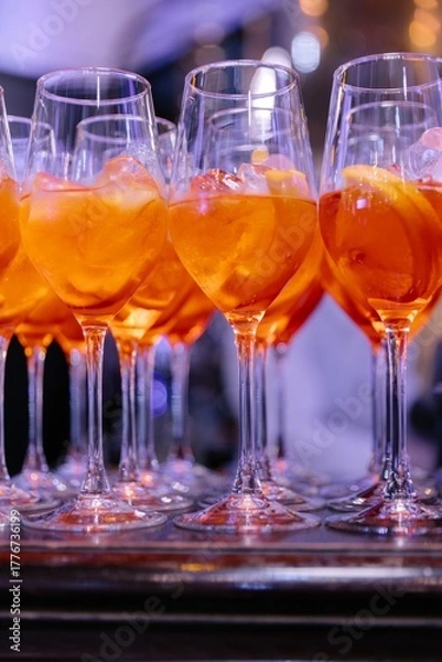 Fototapeta Row of elegant wine glasses filled with bright orange Aperol Spritz cocktails with ice and orange slices, set on a dark bar counter.