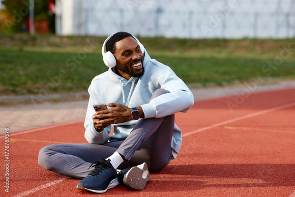 Fototapeta Happy male athlete sits on a running track, smiling while listening to music on his headphones and checking his mobile phone during a workout break.