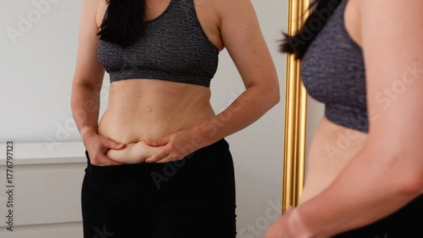 Obraz Woman checking her stomach in front of a mirror, holding belly skin fold. Concept of body image, self-acceptance, fitness progress, postpartum changes, and realistic health goals