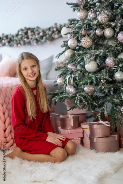 Fototapeta Smiling girl sitting by Christmas tree gifts. A young blonde girl in a red dress sits on a white rug beside a decorated Christmas tree with pink and gold wrapped presents.