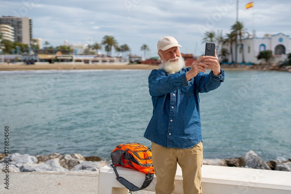 Fototapeta An elderly man with a white beard, wearing a beige cap and denim shirt, takes a selfie with his smartphone by the waterfront, with a Spanish flag, palm trees, and a coastal town in the background