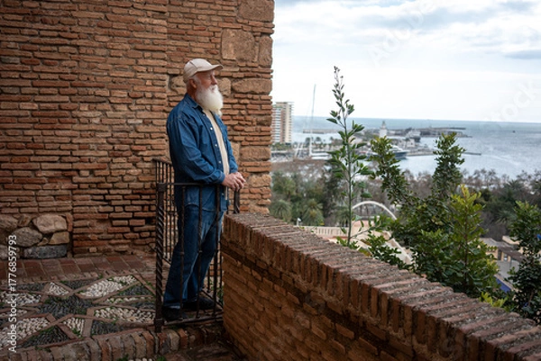 Fototapeta A senior man in a denim jacket leaning on a railing, overlooking the harbor and city skyline, with a scenic view of the coastline and palm trees