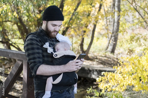 Fototapeta Millennial Dad  with Baby in Carrier Outside Talking and Texting on a Beautiful Fall Day