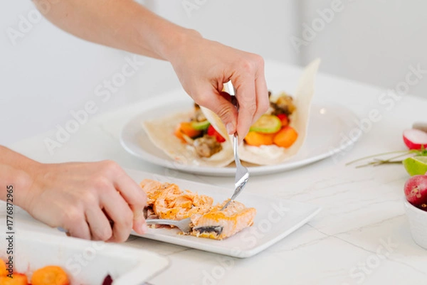 Fototapeta Hands preparing healthy fish tacos for lunch, flaking baked salmon and adding toppings to tortillas