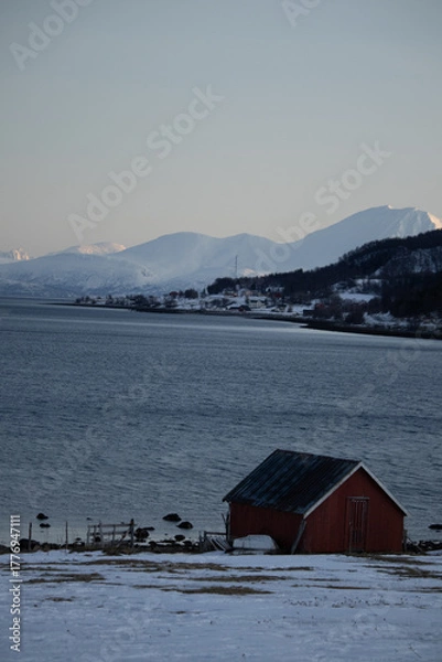 Fototapeta Red cabin by snowy fjord with mountain backdrop in Tromsø, Norway. Arctic winter landscape.