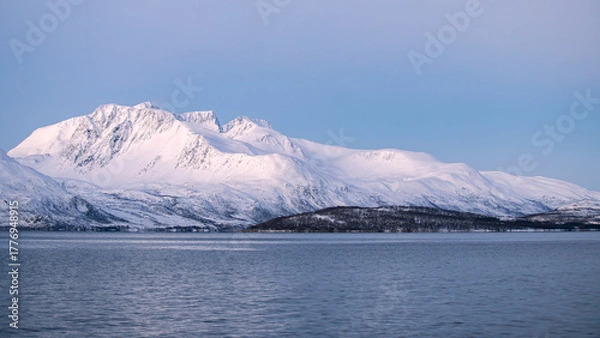 Fototapeta A stunning winter view of a fjord near Tromsø, Norway, featuring calm blue water and towering snow-covered mountains bathed in soft Arctic light.